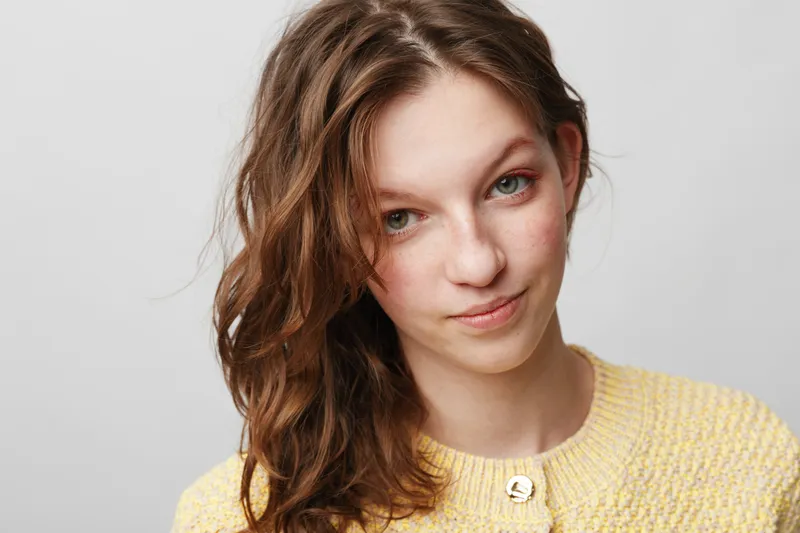 Young woman with wavy brown hair and freckles wears yellow sweater, looking at camera with slight smile. Studio portrait on white background, natural light, casual style and confident mood.