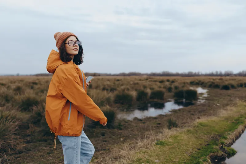 Woman lifestyle outdoors nature river landscape jacket walking autumn travel portrait of a woman in orange jacket with glasses walking near grassy river path under cloudy sky quiet