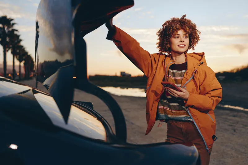 Woman lifestyle outdoors at sunset beside a car, curly hair, warm smile, casual jacket, relaxed confident pose, soft evening light, urban street background, cinematic mood, natural colors