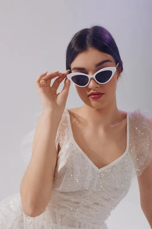 Young woman in a white sequin dress wearing stylish white sunglasses. She poses confidently against a neutral background. Concept of beauty, fashion, and elegance.