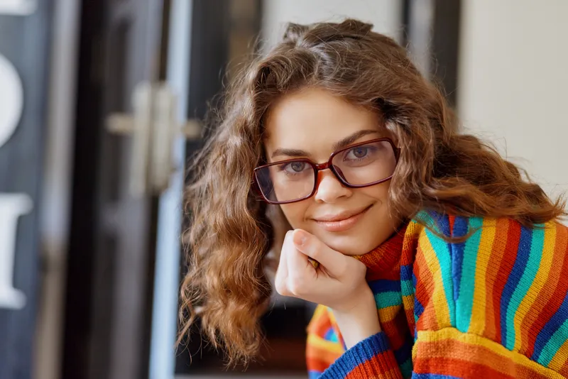 A thoughtful woman with glasses rests her chin on her hand in a bright home setting, wearing a colorful striped sweater. A casual lifestyle portrait emphasizing comfort and creativity.