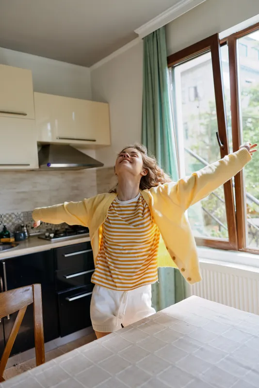 Joyful woman in a yellow jacket stretches arms in a bright kitchen at home, striped tee and white shorts, morning light, open window, carefree mood, candid lifestyle
