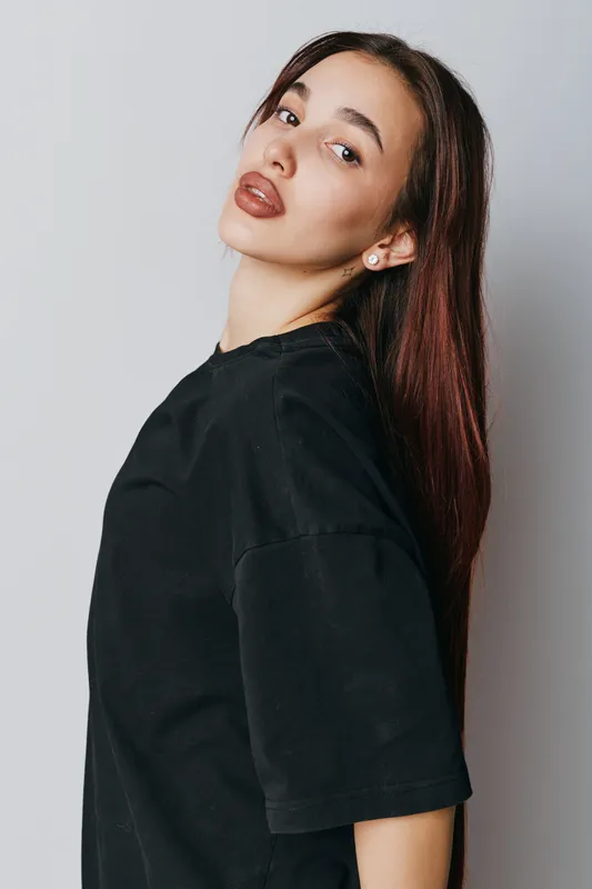 Young woman with long brown hair wearing black t shirt standing against light background looking at camera with confident expression and natural makeup in studio portrait.