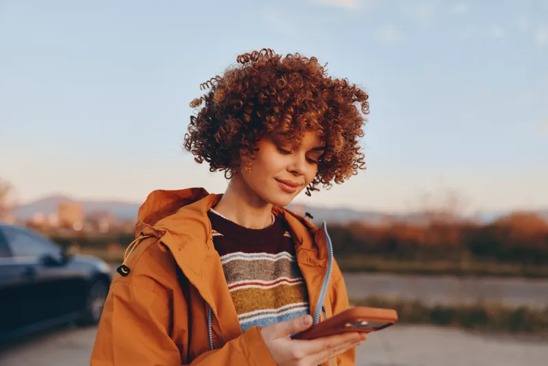 Woman lifestyle outdoors, curly haired woman in orange jacket holding smartphone, smiling gently at sunset, walking near parking and trees, warm natural light, calm relaxed mood