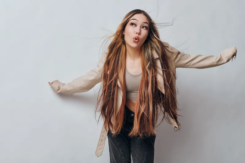 Young woman with long braided hair wearing casual jacket and top posing with arms outstretched making playful expression against plain gray background indoors.