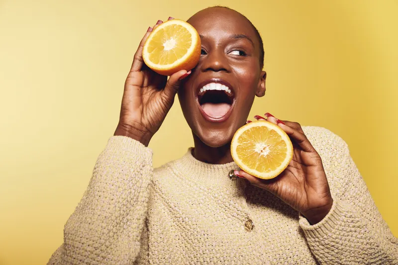 Happy African woman with dark skin holds two halves of a fresh orange fruit near her face, smiling widely. Yellow background and cozy beige sweater create a bright, cheerful mood.