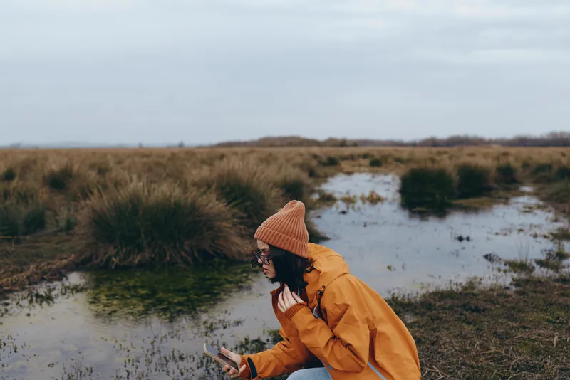 Woman lifestyle outdoors winter countryside, seated on muddy grass by a puddle, wearing brown coat and knit beanie, calm portrait, natural landscape, copy space.