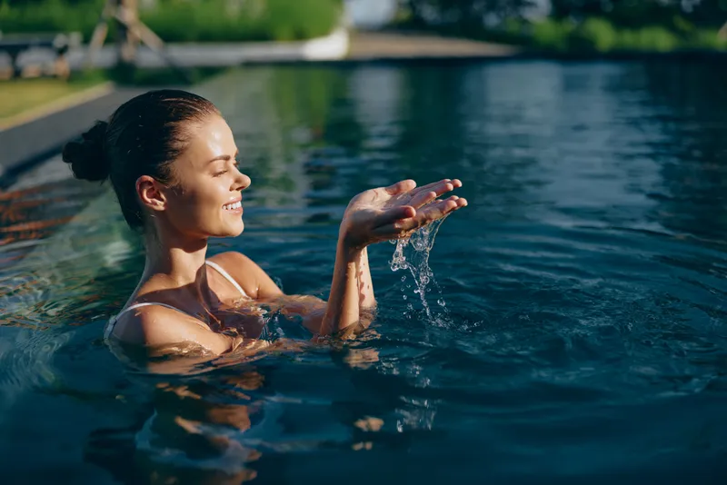 A woman in a pool enjoys the sun, hands cupped as water pours between her fingertips. A moment of simple leisure, calm, and mindfulness in a peaceful outdoor setting.