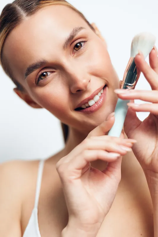 A young woman applies facial cream from a tube, smiling softly as she demonstrates a skincare routine in a well-lit bathroom.