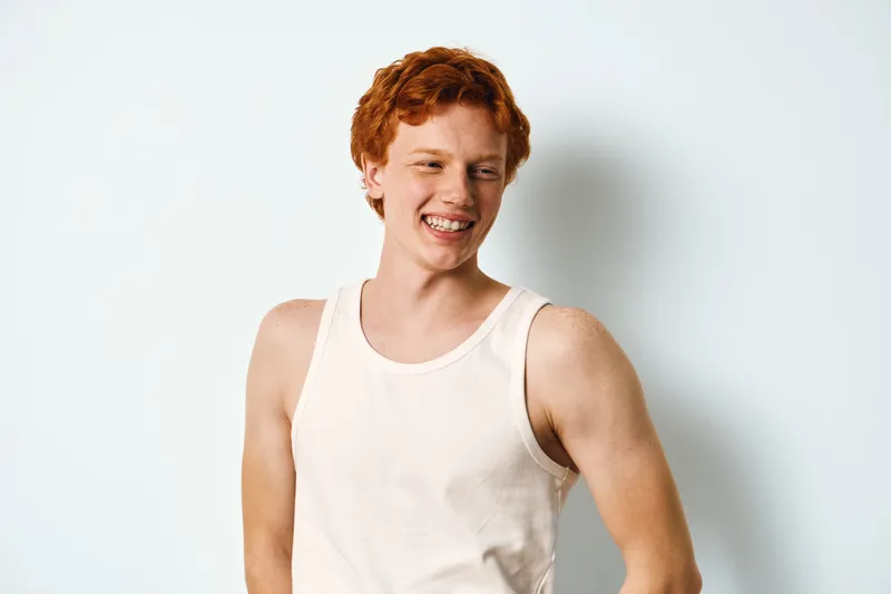 Teenage boy with red hair smiling and laughing wearing white tank top standing against white background in studio. Youth expression, casual style and happiness captured closeup.