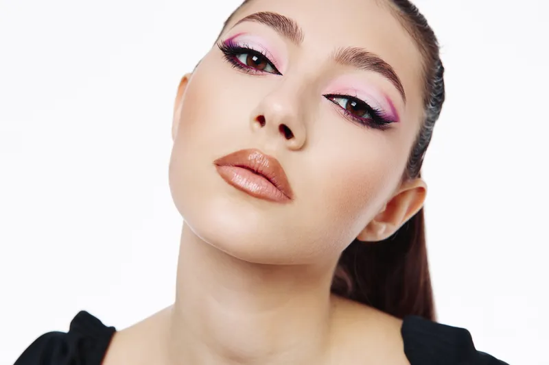 Young woman with creative pink and purple eyeshadow and glossy nude lips poses against white background. Close-up portrait with sleek hair and flawless skin. Fashion beauty concept.