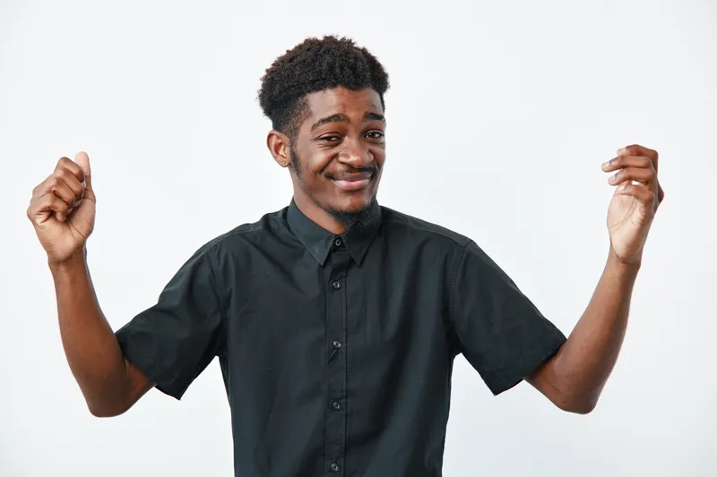 Young black man with dark skin and curly hair wearing black shirt smiling and gesturing with hands on white background. Portrait of confident african ethnicity male.
