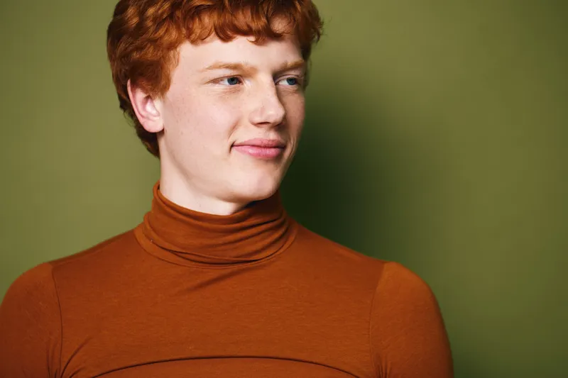 Young man with red hair and freckles wearing brown turtleneck sweater poses sideways against olive green background with slight smile and relaxed expression on face.