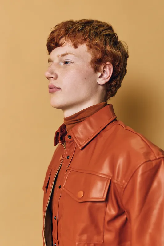Young man with red hair wearing brown leather jacket and turtleneck standing against beige background looking sideways with confident expression and casual style.