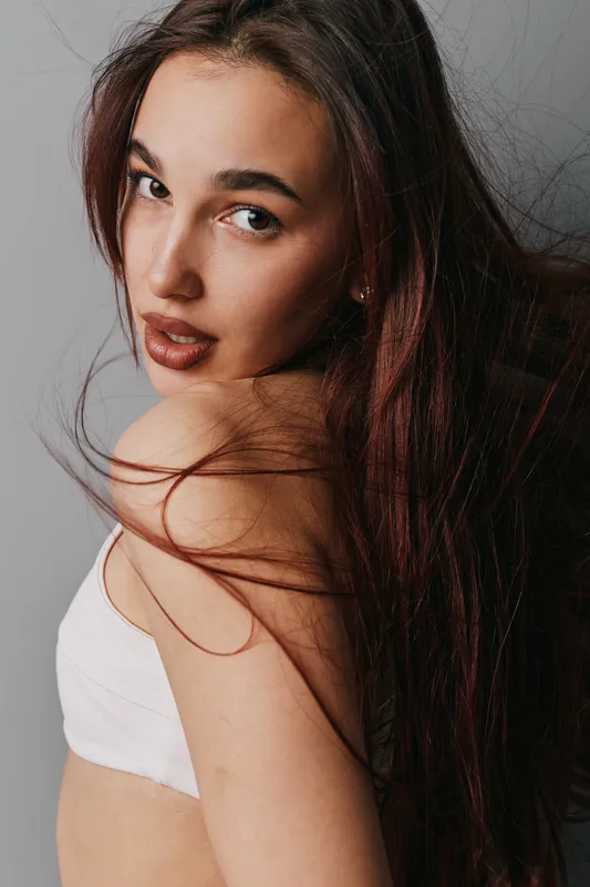 Young woman with long brown hair and natural makeup wearing a white top posing with bare shoulder against dark background. Beauty, portrait and confident look in studio lighting.