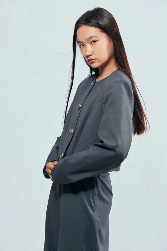 Young woman with long dark hair and casual gray outfit standing against light background, posing with hands in pockets, looking down with neutral expression.