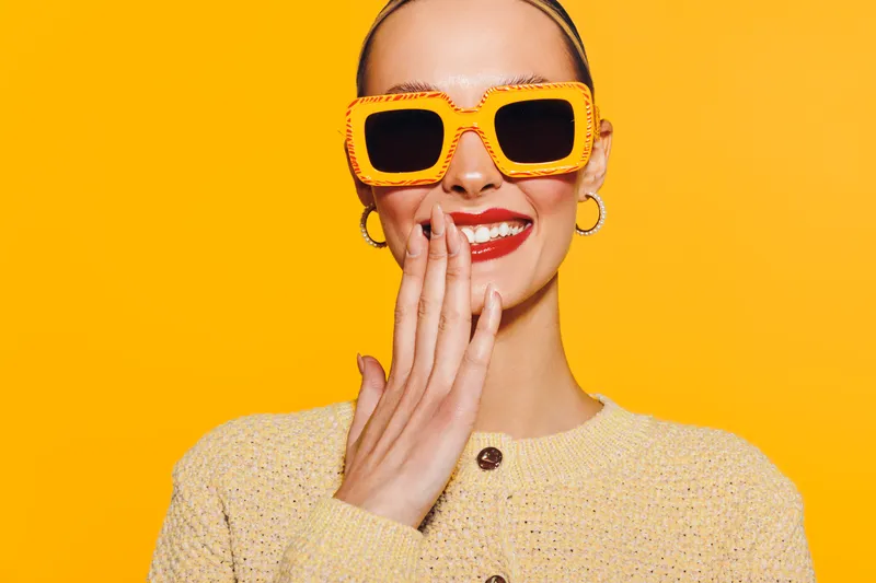 Smiling woman in yellow sunglasses and beige sweater covers mouth with hand on bright yellow background. Concept of happiness, fashion, summer, and positive emotions.