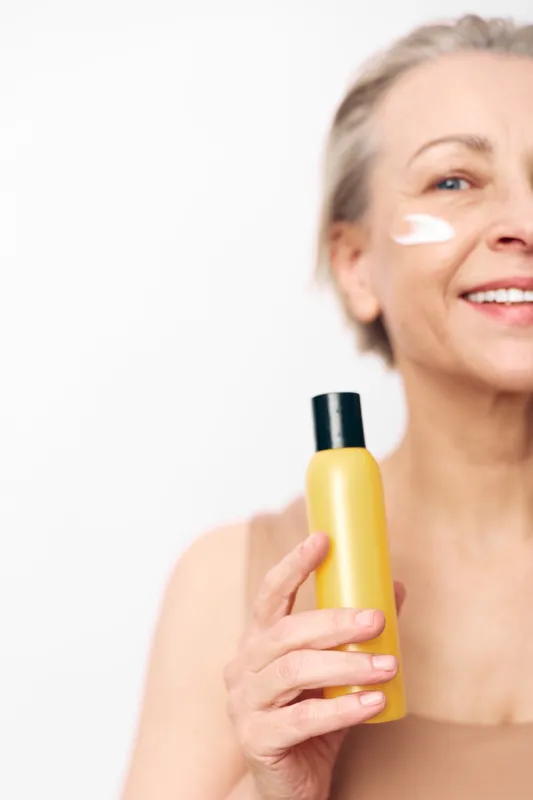 Mature woman with natural skin applying cream on cheek holds yellow bottle. Skincare routine concept with healthy aging, beauty care, and self-confidence on white background.