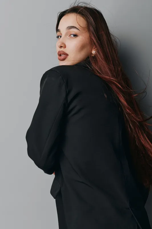 Young woman with long hair wearing black jacket looking over shoulder against gray background. Portrait of confident female model with natural makeup and relaxed pose in studio setting.