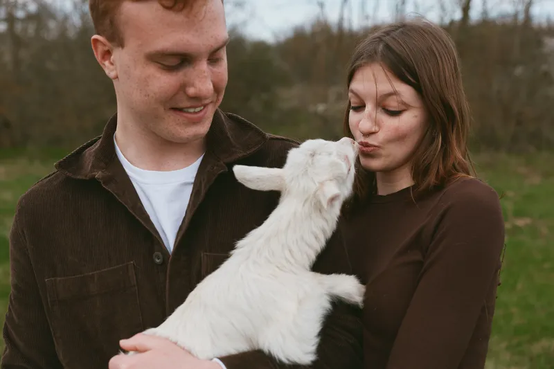 Candid lifestyle shot of a couple with a playful goat kid outdoors, the pair share affection as the animal nuzzles them, rustic field setting, relaxed weekend moment.