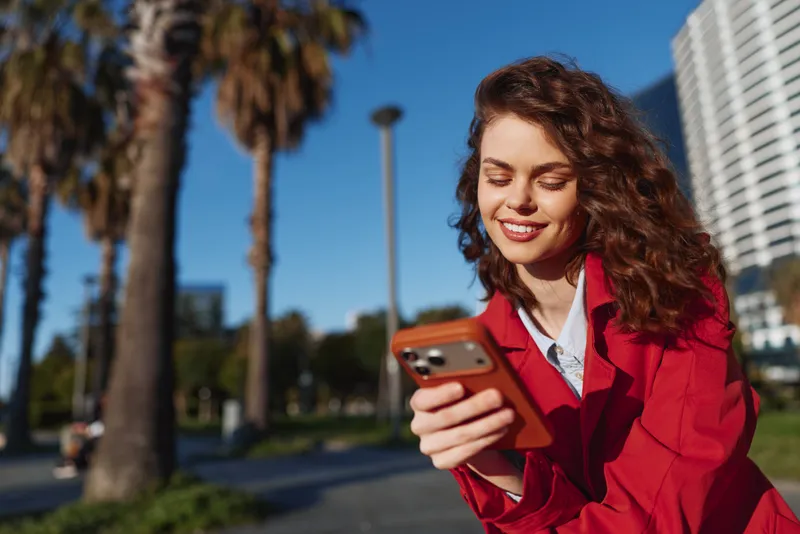 Smiling woman in a red blazer using a smartphone outdoors, sunny day in an urban park, casual fashion, warm, candid moment. Modern style and outdoor vibe.