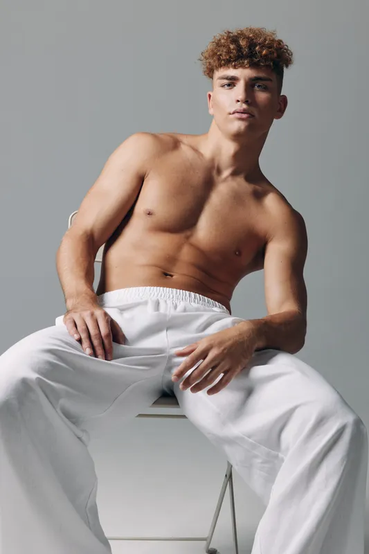 Young man with curly hair sitting shirtless on chair wearing white pants against gray background. Casual pose, confident expression, studio portrait with soft lighting and minimalistic style.