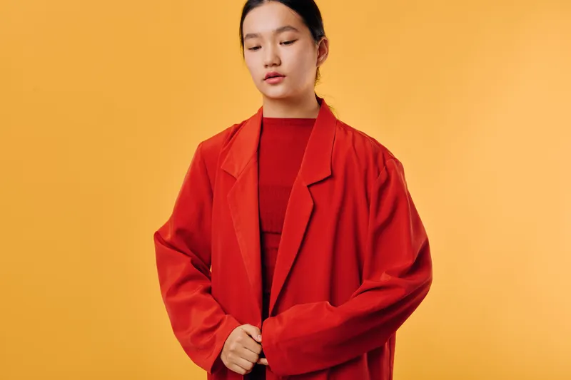 Young woman wearing red oversized jacket and matching top standing against yellow background with closed eyes and calm expression. Portrait of Asian female in casual fashion style studio shot.