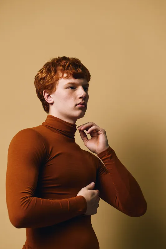 Young man with red hair wearing brown turtleneck sweater posing with thoughtful expression against neutral background in studio portrait. Side view, fashion and style concept.