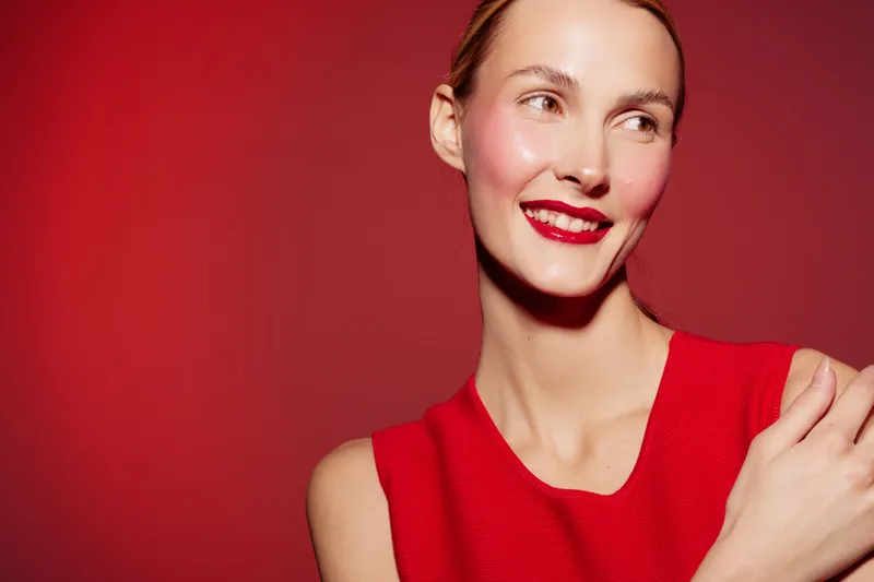 Smiling woman with red lipstick and red sleeveless dress looks away on red background. Portrait of happy female model with natural skin and bright makeup. Concept of beauty and fashion.