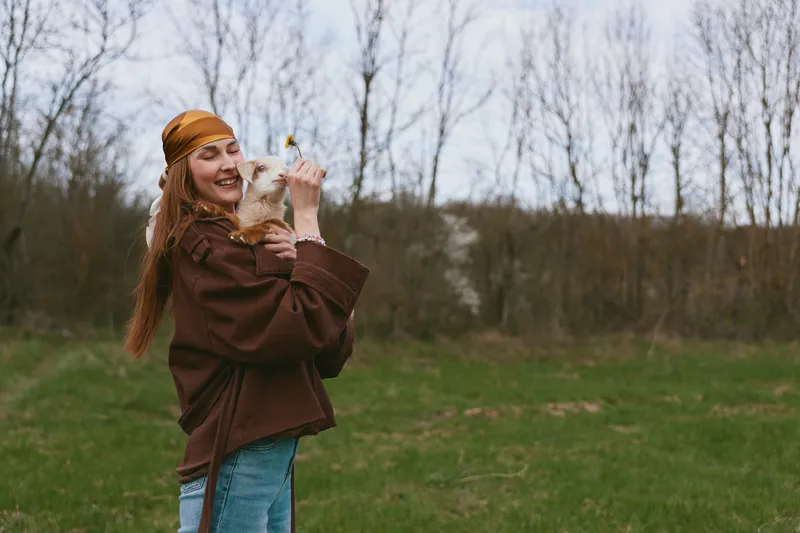 Lifestyle woman outdoors with a goat kid in a field, sharing a playful moment, showing affection and joy against a rustic countryside backdrop.