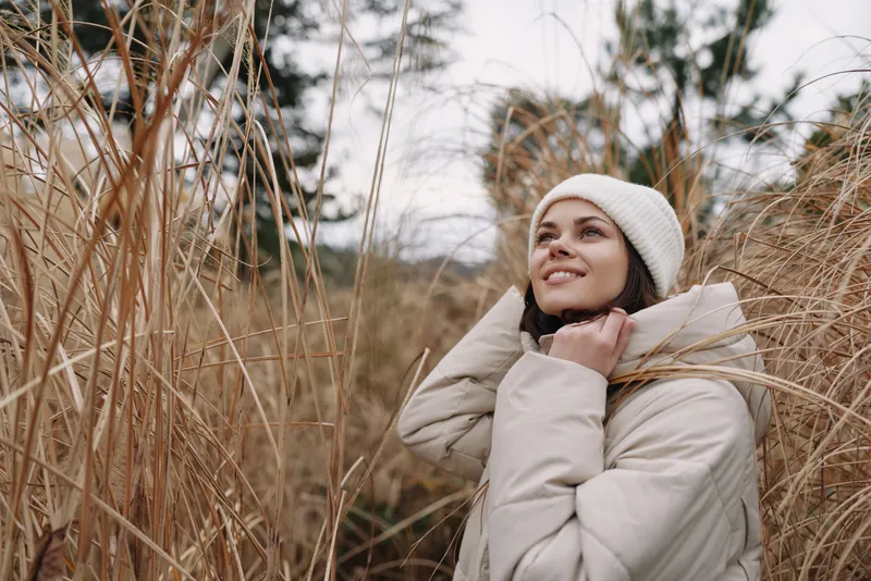 Woman in a warm coat and beanie gazes upward among tall grasses, outdoor portrait in a chilly season, serene mood, natural textures and soft light.