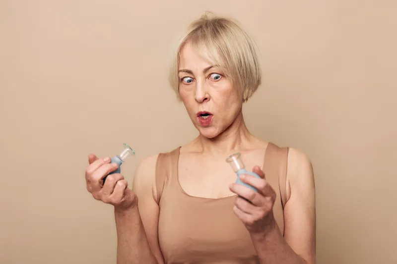 Surprised mature woman in beige top holds two small bottles, comparing contents with curious expression on neutral background. Concept for choice, decision, and skincare product testing.
