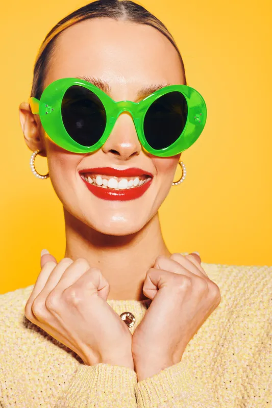 Smiling woman with bright red lipstick wearing large green sunglasses and yellow sweater poses against yellow background. Concept of fashion, summer style, and joyful mood.
