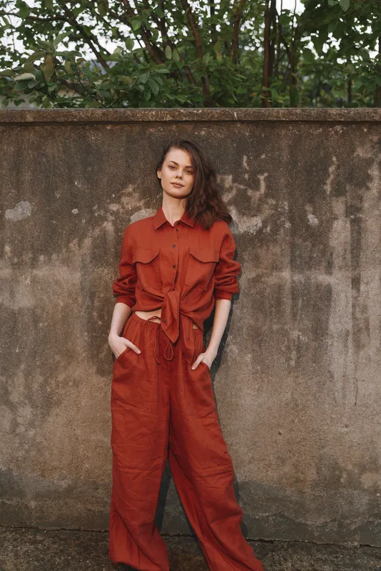 Young woman in a rust jumpsuit stands against a weathered wall with hands in pockets, radiating casual confidence and urban street style