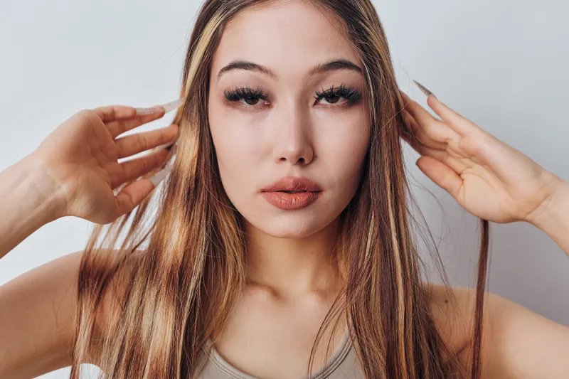 Woman with long hair and natural makeup posing with hands near head, looking directly at camera with calm expression and soft lighting on face.