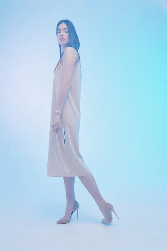 Elegant woman in a beige dress and high heels poses in a soft blue studio light. Her long hair flows naturally, showcasing beauty and fashion concepts with a calm expression.