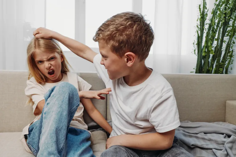 Two children sitting on couch, boy pulling girl’s hair, girl upset and resisting indoors, family conflict and sibling dispute concept.