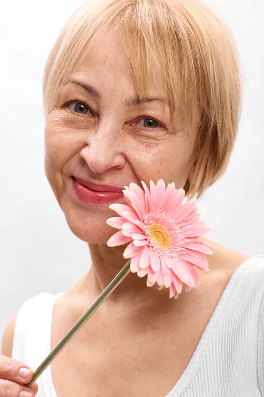 Woman portrait closeup with pink daisy flower near face, gentle smile and soft lighting, short blonde hair, clean white background, beauty and skincare concept, calm wellness mood