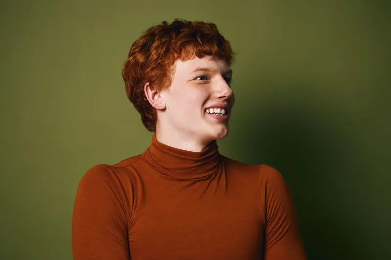 Young redhead man smiling and looking sideways wearing brown turtleneck sweater on green background in studio portrait with natural light and casual style.