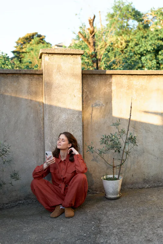 Woman sits on the ground against a sunlit wall, wearing a red jumpsuit, holding a smartphone, capturing a relaxed outdoor lifestyle moment beside a potted plant