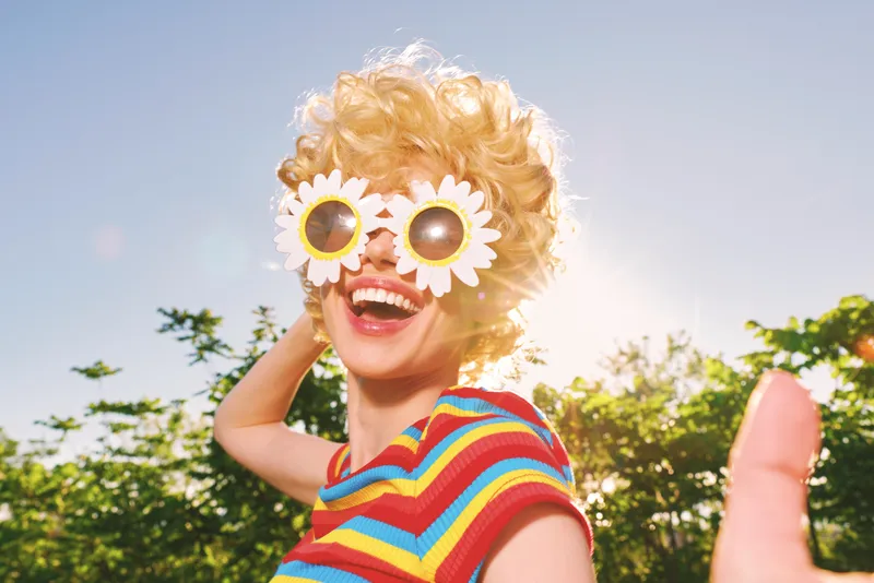 Cheerful woman outdoors, wearing colorful striped top and flower sunglasses, laughing under sunlit sky, relaxed pose, vibrant retro vibe, joyful moment, bright greenery backdrop