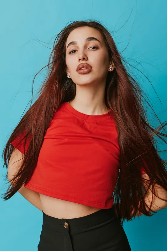 Young woman with long hair wearing red crop top and black pants posing confidently against blue background. Portrait of female model with natural makeup and earrings in studio.