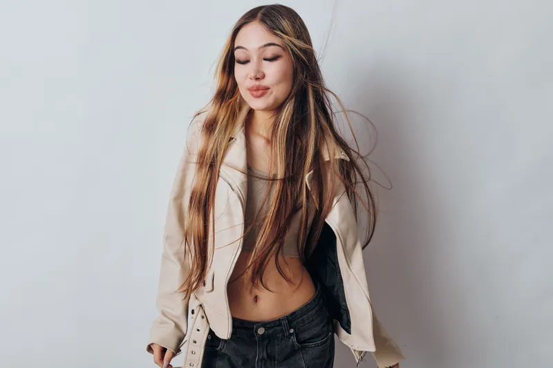 Young woman with long hair wearing casual beige jacket and black pants posing with closed eyes, relaxed expression and minimal makeup on light background indoors in studio.