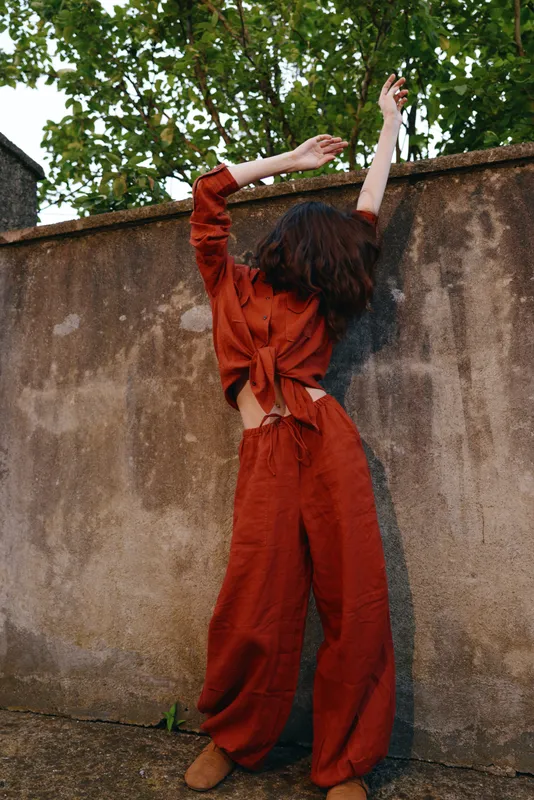 Young woman with a back view reaches up against a textured wall outdoors in a loose orange jumpsuit, capturing a candid moment of freedom and quiet confidence