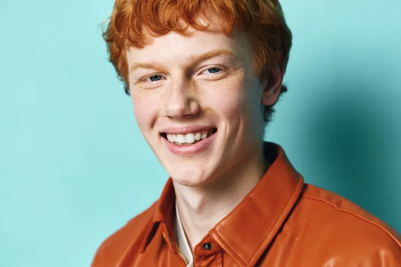 Young man with red hair smiling and wearing orange shirt on blue background. Portrait of happy male person with casual style and friendly expression in studio photography.
