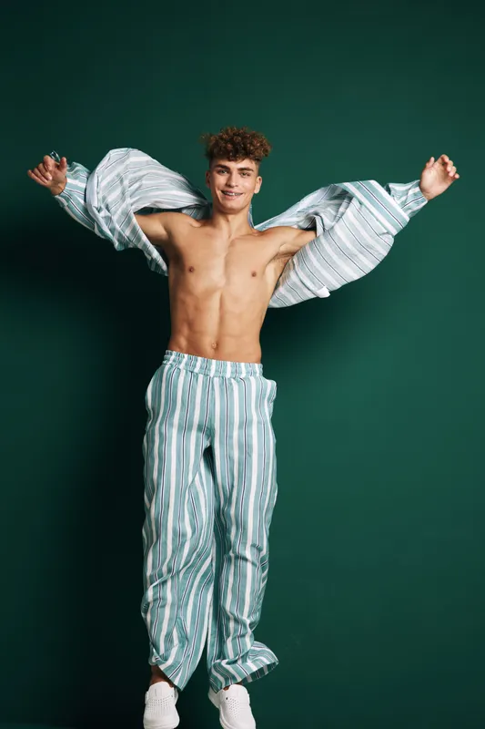 Young man with curly hair wearing striped pants and open shirt jumping against green background. Casual style, smiling, energetic and confident male model in motion with bare chest.