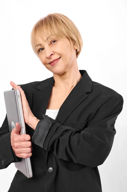 Mature blonde woman portrait, smiling while holding a tablet, wearing a black blazer and white top, studio background, confident business style, clean soft lighting