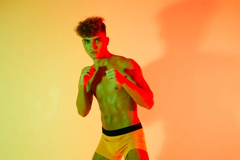 Young athletic man with curly hair posing in fighting stance with fists up against colorful orange and yellow background for fitness and boxing training concept.