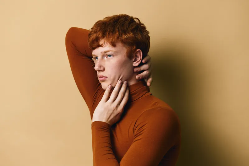 Young man with red hair posing with hand on neck and face wearing brown long sleeve turtleneck sweater against beige background with thoughtful expression.