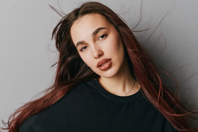 Young woman with long red hair and natural makeup poses against gray background wearing black top. Portrait of confident female with loose hair and serious expression in studio light.